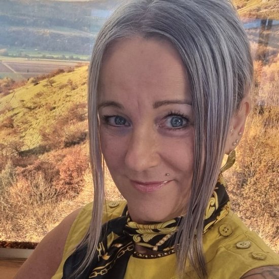 Selbstporträt einer Frau mit silbergrauen Haaren und blauen Augen. Sie trägt ein gelbes Oberteil und einen schwarz-gelben Schal. Ein Tattoo ist auf ihrem linken Oberarm sichtbar. Im Hintergrund ist eine unscharfe Hügellandschaft zu erkennen.