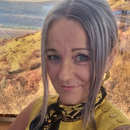 Selbstporträt einer Frau mit silbergrauen Haaren und blauen Augen. Sie trägt ein gelbes Oberteil und einen schwarz-gelben Schal. Ein Tattoo ist auf ihrem linken Oberarm sichtbar. Im Hintergrund ist eine unscharfe Hügellandschaft zu erkennen.