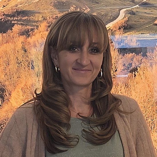 Eine Frau mit braunen Haaren und Pony lächelt in die Kamera. Sie trägt eine beige Strickjacke und Jeans. Im Hintergrund ist eine Landschaftsmalerei mit einem Hügel und einer Burgruine zu erkennen.