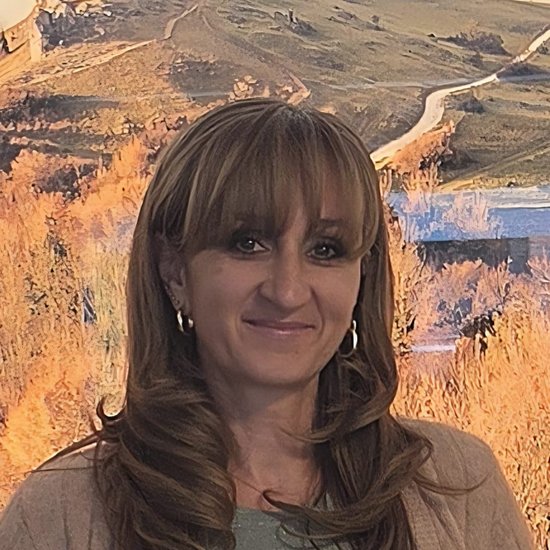 Eine Frau mit braunen Haaren und Pony lächelt in die Kamera. Sie trägt eine beige Strickjacke und Jeans. Im Hintergrund ist eine Landschaftsmalerei mit einem Hügel und einer Burgruine zu erkennen.