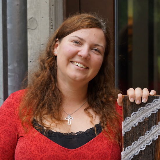 Eine lächelnde Frau mit lockigen, braunen Haaren und einem roten Oberteil hält ein dunkles Holzbrett, an dem zwei Streifen weißer Spitze befestigt sind.