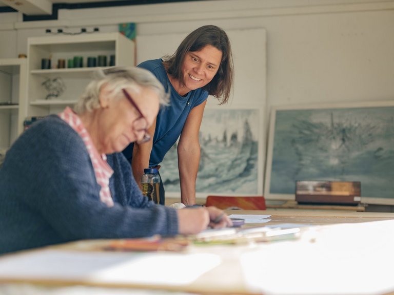 Eine Frau schaut einer anderen Frau dabei über die Schulter, wie sie an einem Tisch etwas malt.