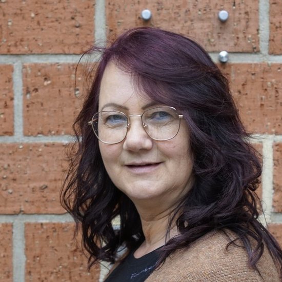 Eine Frau mit dunklen, gewellten Haaren und einer Brille steht lächelnd vor einer roten Backsteinmauer. Sie trägt eine braune Strickjacke über einem schwarzen Oberteil.