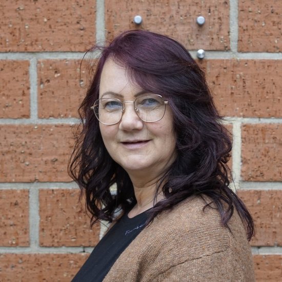 Eine Frau mit dunklen, gewellten Haaren und einer Brille steht lächelnd vor einer roten Backsteinmauer. Sie trägt eine braune Strickjacke über einem schwarzen Oberteil.