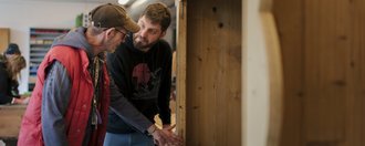 Zwei Menschen stehen in einer Schreinerwerkstatt und begutachten gemeinsam ein Werkstück.