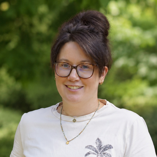 Portrait einer lächelnden Frau mit Brille und dunklem Haar im Dutt. Sie trägt ein weißes T-Shirt mit einem kleinen Palmendruck und steht vor einem unscharfen, grünen Naturhintergrund.