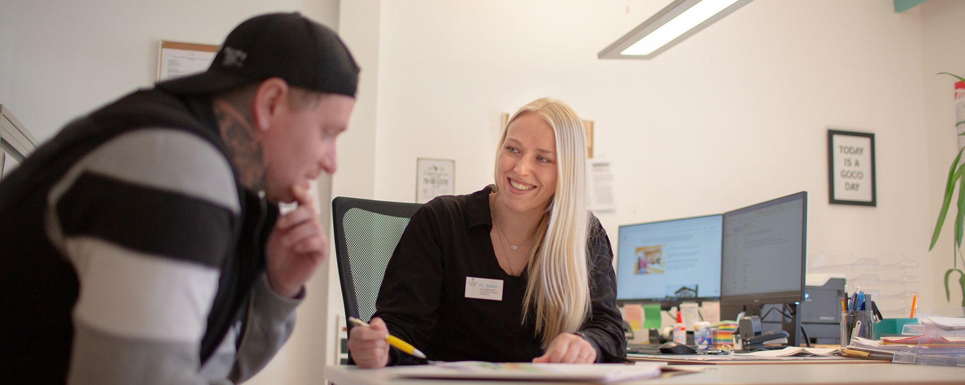 im Büro: eine freundliche Mitarbeiterin erklärt einem Besucher etwas anhand eines Schriftstücks