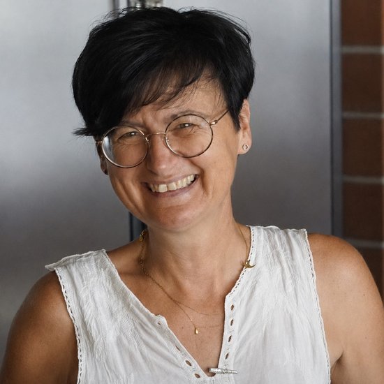 Porträt einer lächelnden Frau mit kurzen schwarzen Haaren und einer runden Brille. Sie trägt ein weißes, ärmelloses Oberteil und blickt freundlich in die Kamera.
