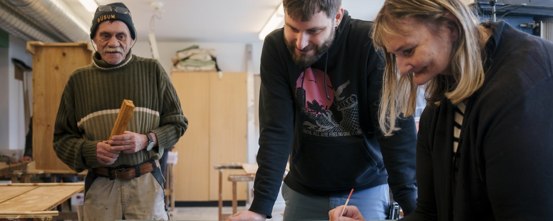 Eine Person bemalt eine Holztür in einer Schreinerwerkstatt, während zwei andere Personen interessiert dabei zusehen.