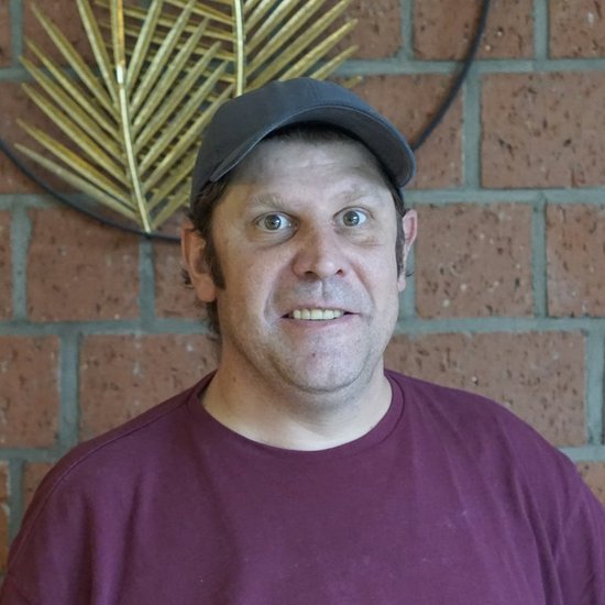 Ein lächelnder Mann mit einem dunklen Cap und einem weinroten T-Shirt steht vor einer roten Backsteinwand, an der eine dekorative Metallpflanze hängt.