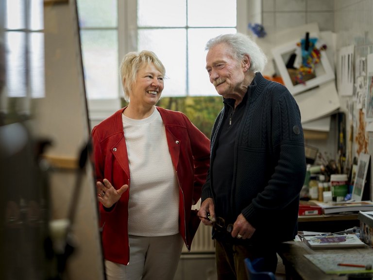 Eine Frau und ein Mann stehen vor einer Leinwand in einer Künstlerwerkstatt und diskutieren das Werk des Mannes.