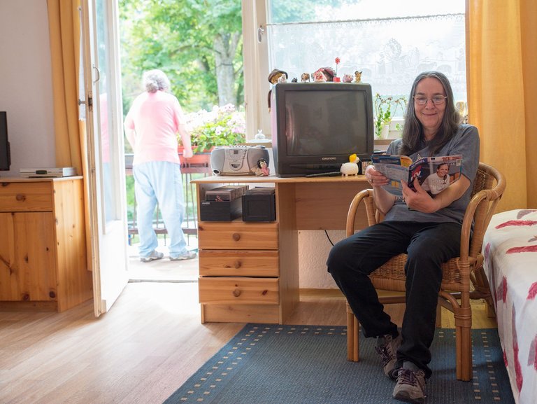 eine Frau sitzt in ihrem Zimmer, blättert in einer TV-Zeitschrift, während im Hintergrund eine weitere am Balkon steht 