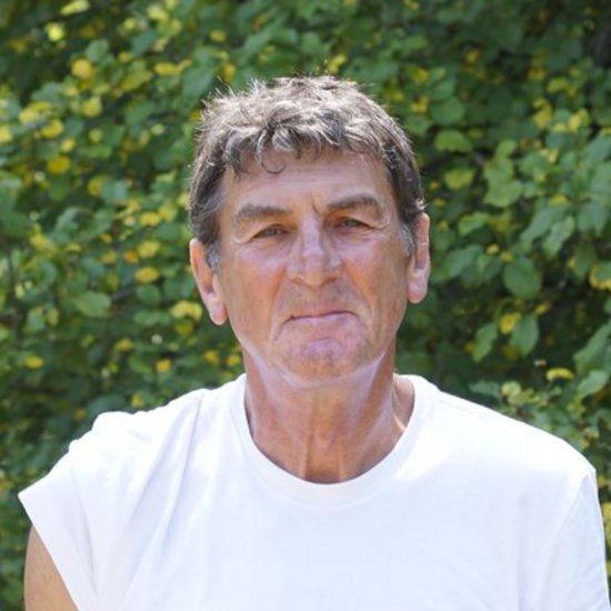 Ein älterer Mann mit grauen Haaren und einem weißen ärmellosen T-Shirt steht vor einem Hintergrund aus grünen Bäumen und blickt in die Kamera.