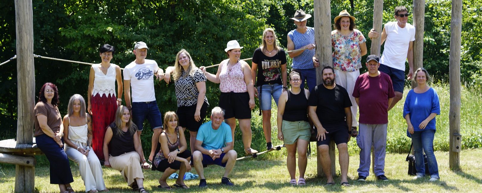 Eine Gruppe von etwa 16 Erwachsenen posiert lächelnd im Sommer auf einer Wiese. Im Hintergrund sind hohe Holzpfosten und Bäume zu sehen. Die Personen stehen oder hocken in Reihen und blicken in die Kamera.